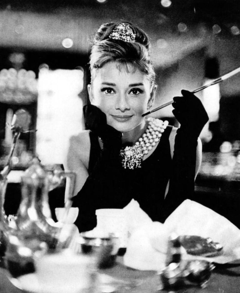 K šatům v podání Audrey neodmyslitelně patří také doplňky – dlouhé černé rukavice, diamantová korunka, perleťový náhrdelník, cigaretová špička a velký černý klobouk.